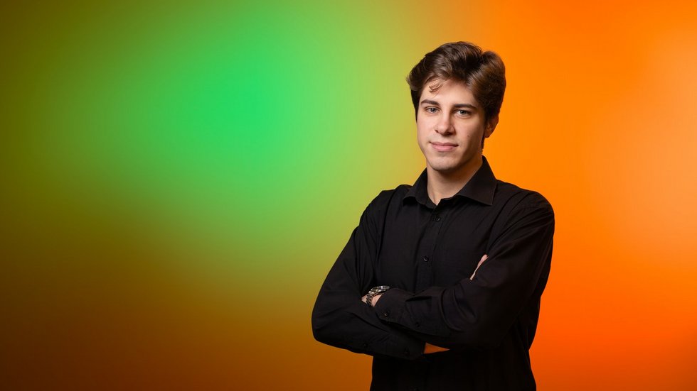 Wysoki chłopak, szatyn ubrany w czarna koszulę stoi z założonymi rękami na tle pomarańczowo zielonej ściany