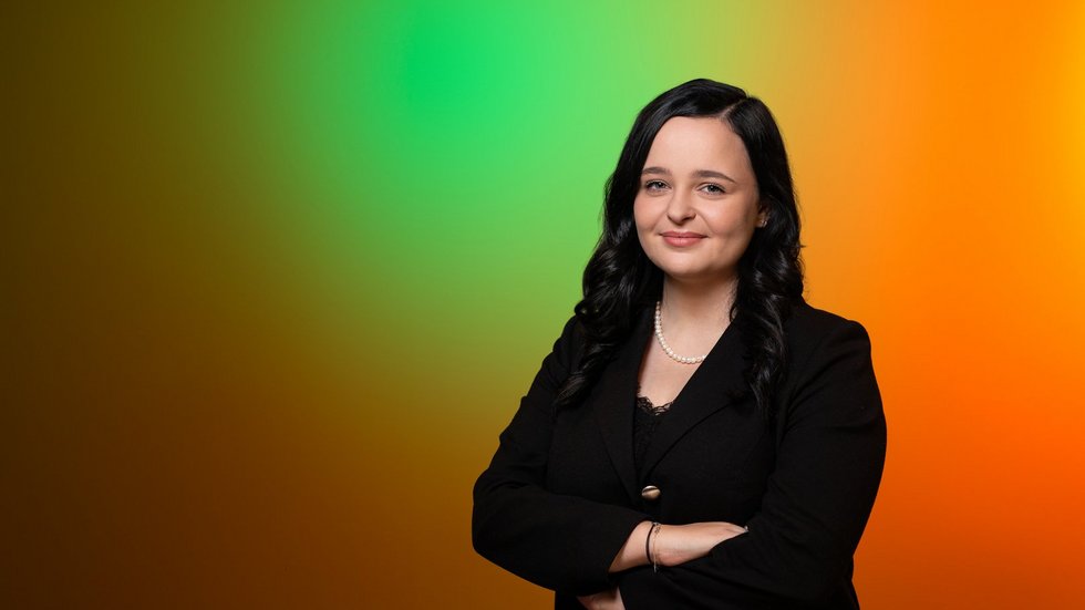 Młoda dziewczyna z długimi, ciemnymi włosami stoi z założonymi rękami na kolorowym , zielono-pomarańczowym tle.